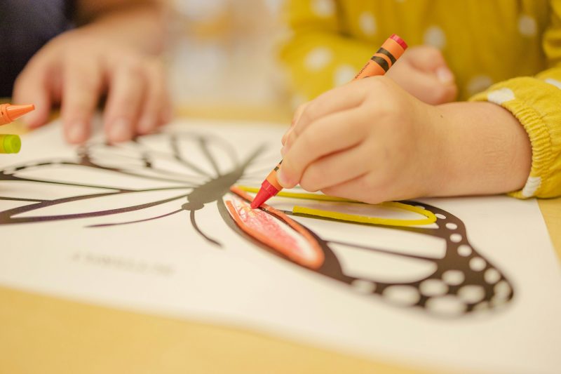 Kinderhände malen mit einem roten Wachsmalstift eine Schmetterlingszeichnung aus