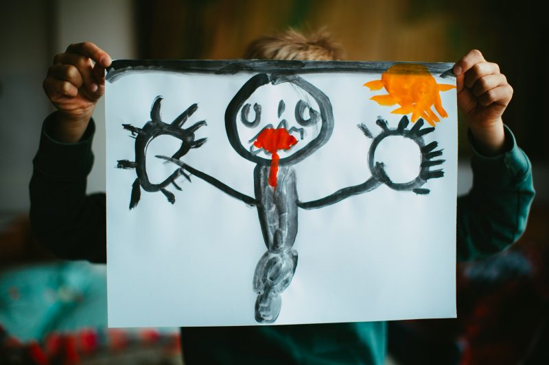 Ein Kind hält ein selbst gemaltes Bild von einer Figur mit Sonne hoch, sein Gesicht ist hinter dem Blatt verborgen