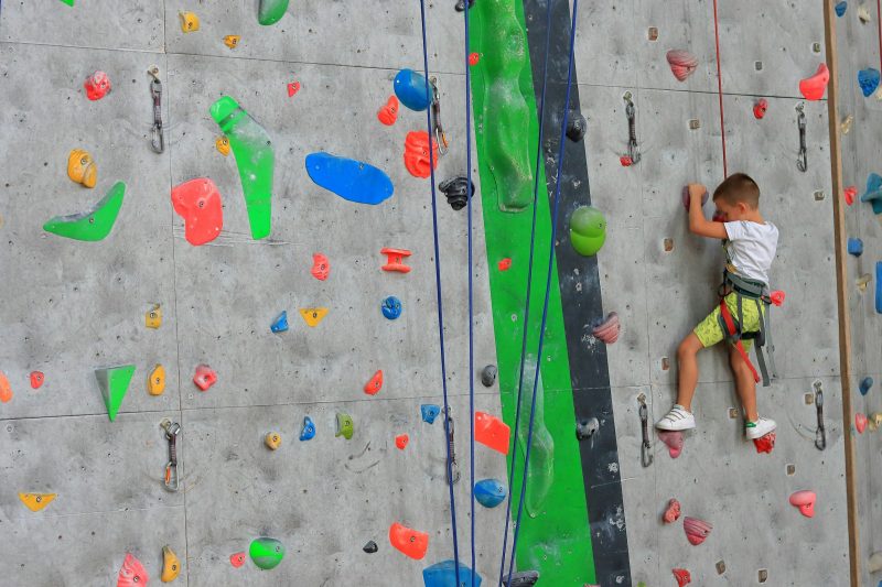 Ein Junge klettert gesichert an einer bunten Kletterwand nach oben