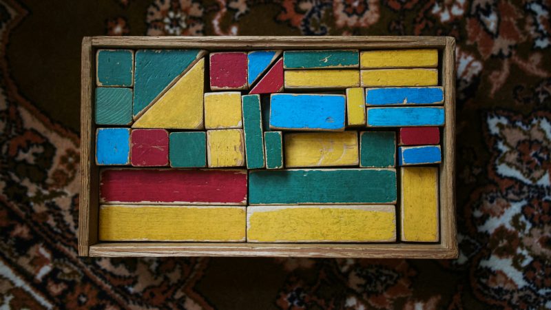 Eine Holzkiste mit bunten, ordentlich eingeräumten Bauklötzen steht auf einem gemusterten Teppich