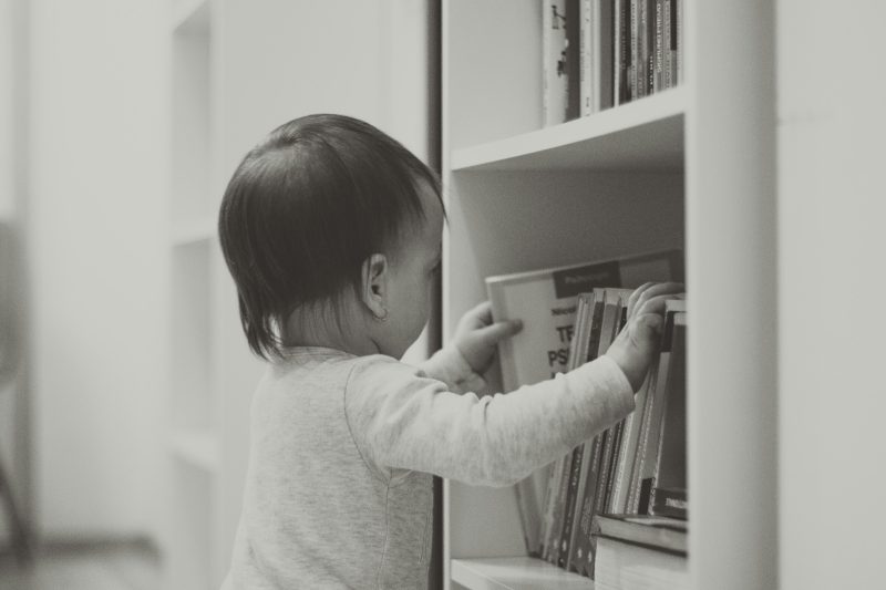 Ein Kleinkind stellt von hinten gesehen ein Buch in ein weißes Bücherregal, Schwarz-Weiß-Foto