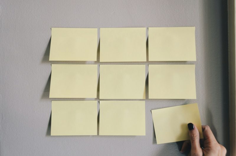 Eine Hand hängt einen von mehreren leeren gelben Klebezetteln an eine helle Wand