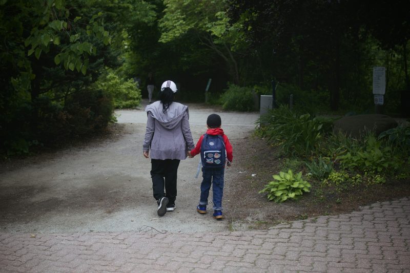 Eine erwachsene Person und ein Kind mit rotem Rucksack gehen von hinten gesehen Hand in Hand über einen Parkweg