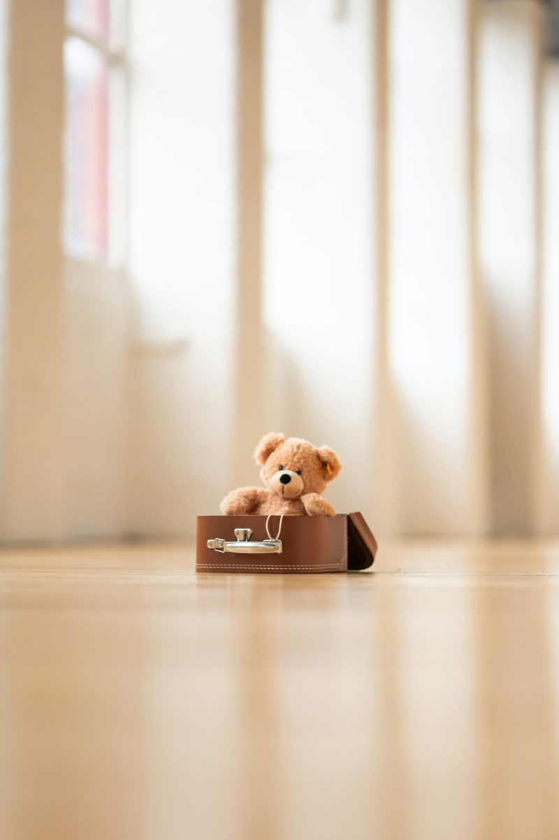 Ein Teddybär sitzt in einem kleinen braunen Koffer, der auf einem hellen Holzboden steht