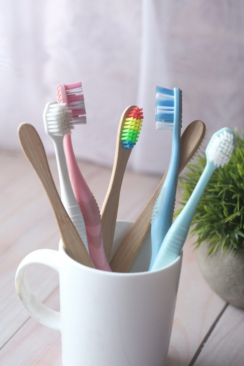 Several colourful plastic and wooden toothbrushes standing in a white ceramic beaker beside a small plant