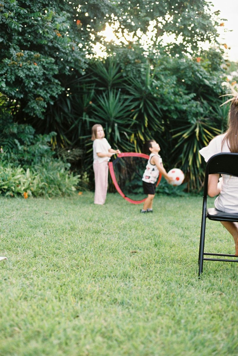 Kinder spielen in einem grünen Garten mit Ball und Reifen, am Bildrand sitzt eine erwachsene Person