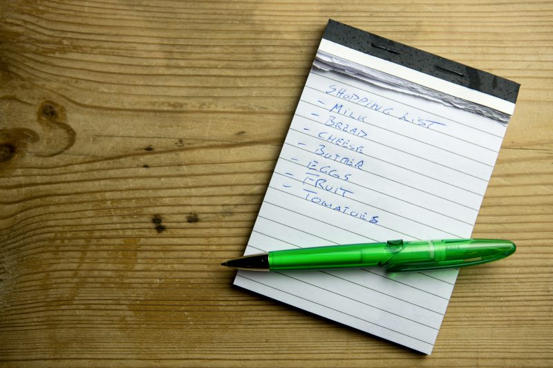 A weekly calendar on a kitchen wall with a handwritten shopping list beside it