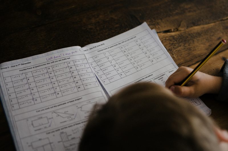 A child sitting at a wooden table over an exercise book, with a tablet showing a task list beside them