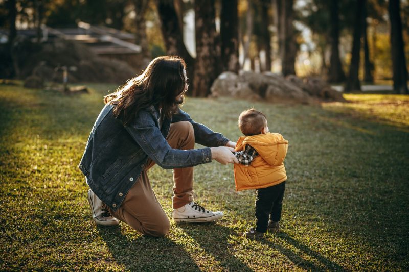 A woman in a denim jacket kneeling in a park at eye level with a toddler in an orange gilet, holding their hands
