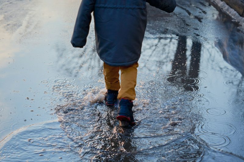 A child in a winter coat and boots, seen from behind, stomping through a large puddle
